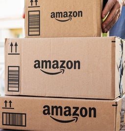 A person picks up stacked cardboard Amazon packages on a wooden table, with a blurred background of a sunny balcony.