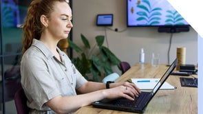 Woman sits in an office working on her laptop with a notebook next to her