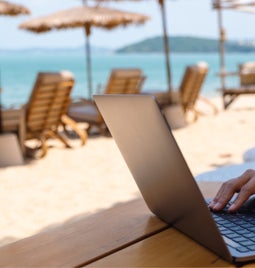 Person working at a laptop on a white sand beach with lounge chairs and umbrellas in the background
