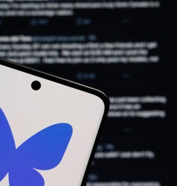 Bluesky logo on a phone, held in front of a screen showing the Bluesky feed