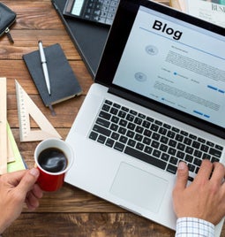 man working on laptop on a wooden table with stationary and paper scattered around him. The laptop is open on a page that says blog