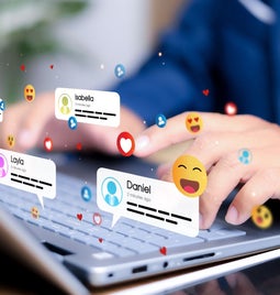 A person types on a laptop keyboard, surrounded by floating social media notifications and emojis representing interactions with friends.
