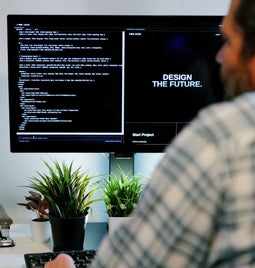 A person sits at a desk, programming on a computer with code on the screen, surrounded by plants and a coffee mug.