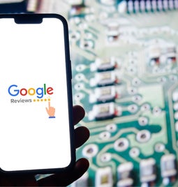 A hand holds a smartphone displaying the Google Reviews logo against a blurred circuit board background.