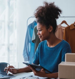 A person in a blue sweater sits at a desk with a laptop, tablet, and stationery, surrounded by clothing and shipping boxes.