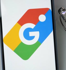 A smartphone displaying the Google logo, positioned beside a white keyboard and a pair of glasses on a gray surface.