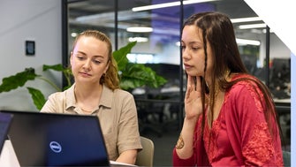 Two professional women collaborating on a project using a laptop in an office setting.