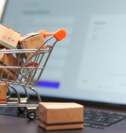 A miniature shopping cart filled with small cardboard boxes rests on a laptop keyboard, with an online shopping interface in the background.