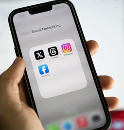 hand holding an iphone open on a homepage folder called social networking, containing X, Threads, Instagram, and Facebook apps