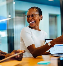 A professional meeting scene with two individuals engaging in discussion, multiple laptops, and documents on a wooden conference table.