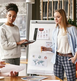 Three professionals collaborate in a modern workspace, discussing plans with a laptop, charts, and diagrams displayed on a board.