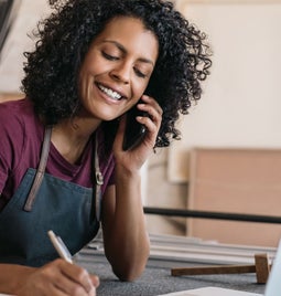 Smiling businesswoman in a t-shirt and apron is on the phone, and makes notes on a pad at her desk, in front of an open laptop