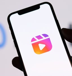 A hand holding a smartphone displaying the Instagram Reels logo, with a blurred Meta logo in the background.