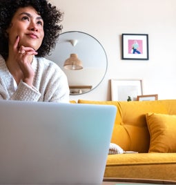 A person in a cozy sweater sits thoughtfully on a yellow couch, focused on a laptop surrounded by a stylish, modern interior.