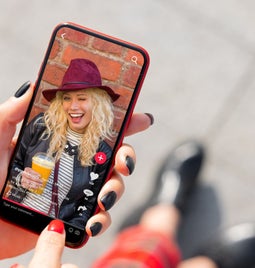 A hand holds a smartphone displaying a social media video, featuring a person with curly hair and a drink, against a brick background.