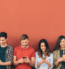 7 people leaning against a red wall looking down at their phones.