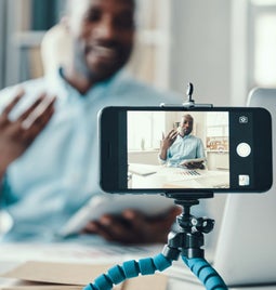 A person gestures while speaking to a smartphone on a tripod, surrounded by office supplies and paperwork in a bright workspace.