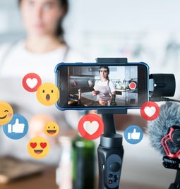A smartphone on a gimbal captures a video in a kitchen, surrounded by animated social media reaction icons.
