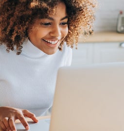 A person with curly hair sits at a kitchen table, focused on a laptop, with a cozy kitchen backdrop.
