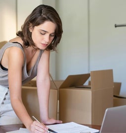 A woman in front of her laptop writing down stock numbers on a notepad