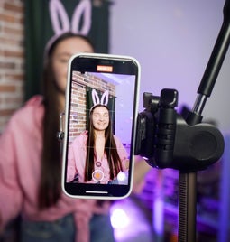 lady in bunny ears and a pink jumper filming herself with a smartphone held up with a tripod