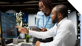 Two colleagues discuss a project at a modern office, focusing on a computer screen. One holds a tablet while pointing at the screen.