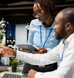 Two colleagues discuss a project at a modern office, focusing on a computer screen. One holds a tablet while pointing at the screen.
