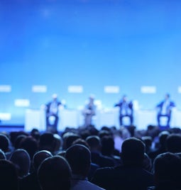 Large audience sat in chairs in front of a stage with speakers sat on chairs and a person stood to the side to moderate
