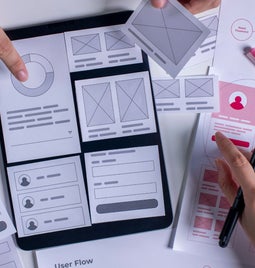 Hands arranging wireframe templates and mockups on a table, surrounded by design tools, illustrating the user interface design process.