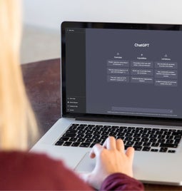 A person with long blonde hair sits at a wooden table, using a laptop displaying the ChatGPT interface with features and limitations.