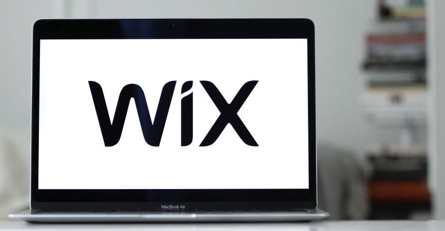 A close-up of a MacBook Air displaying the Wix logo on its screen, with a blurred background of bookshelves.