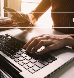 Hands typing on a keyboard while holding a phone with a search bar graphic on top